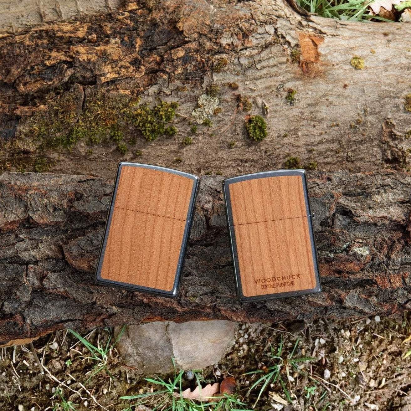 Lifestyle image of WOODCHUCK USA Cherry Emblem Windproof Lighter, laying on a log with one lighter showing the front and the other showing the back