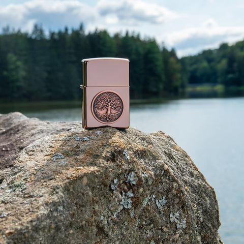 Lifestyle image of Tree of Life Emblem High Polish Rose Gold Windproof Lighter standing on a rock, with the lake and trees in the background.