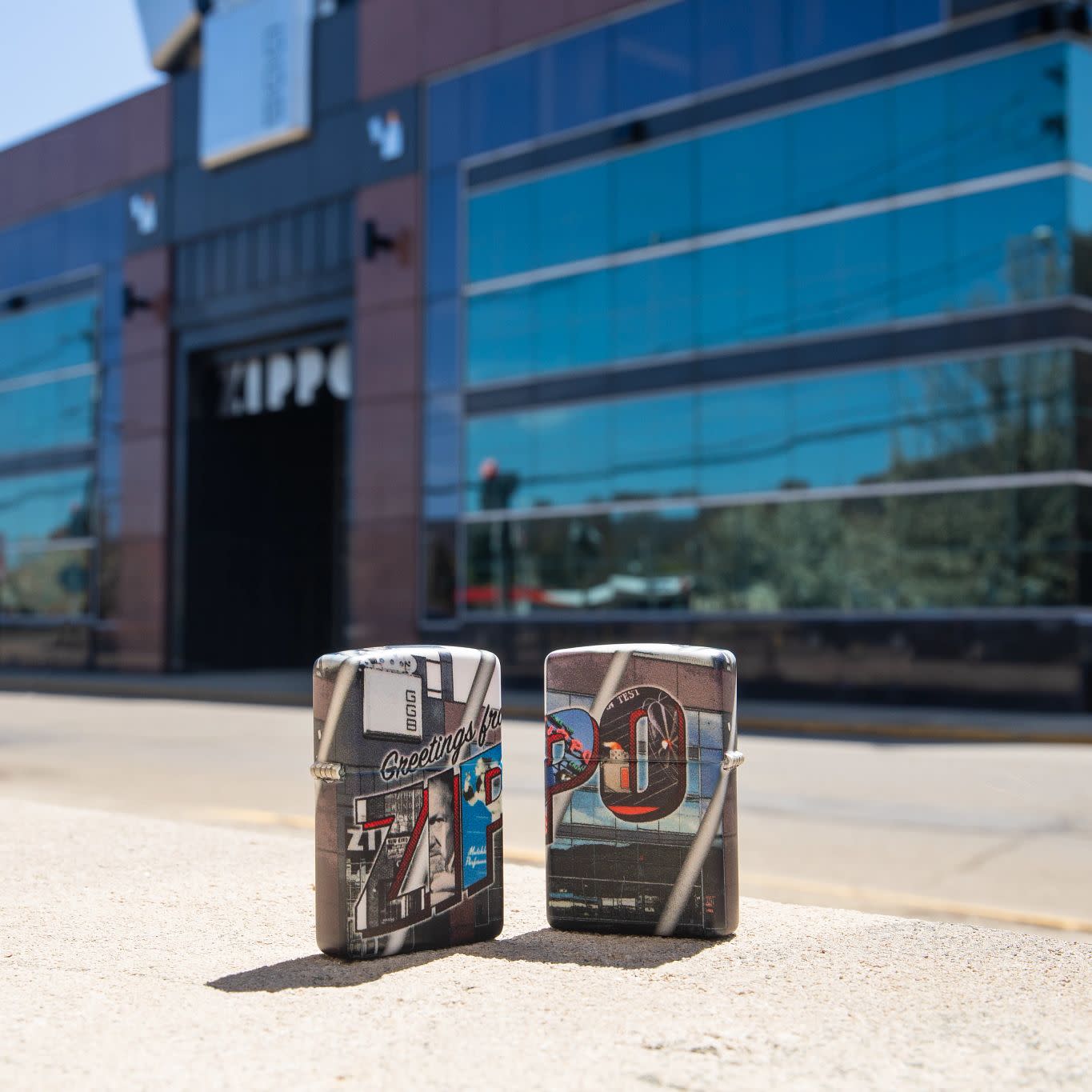 Lifestyle image of two Greetings from Zippo 540 Color Windproof Lighter standing in front of the Zippo headquarters building. One lighter is showing the front of the design, the other is showing the back.